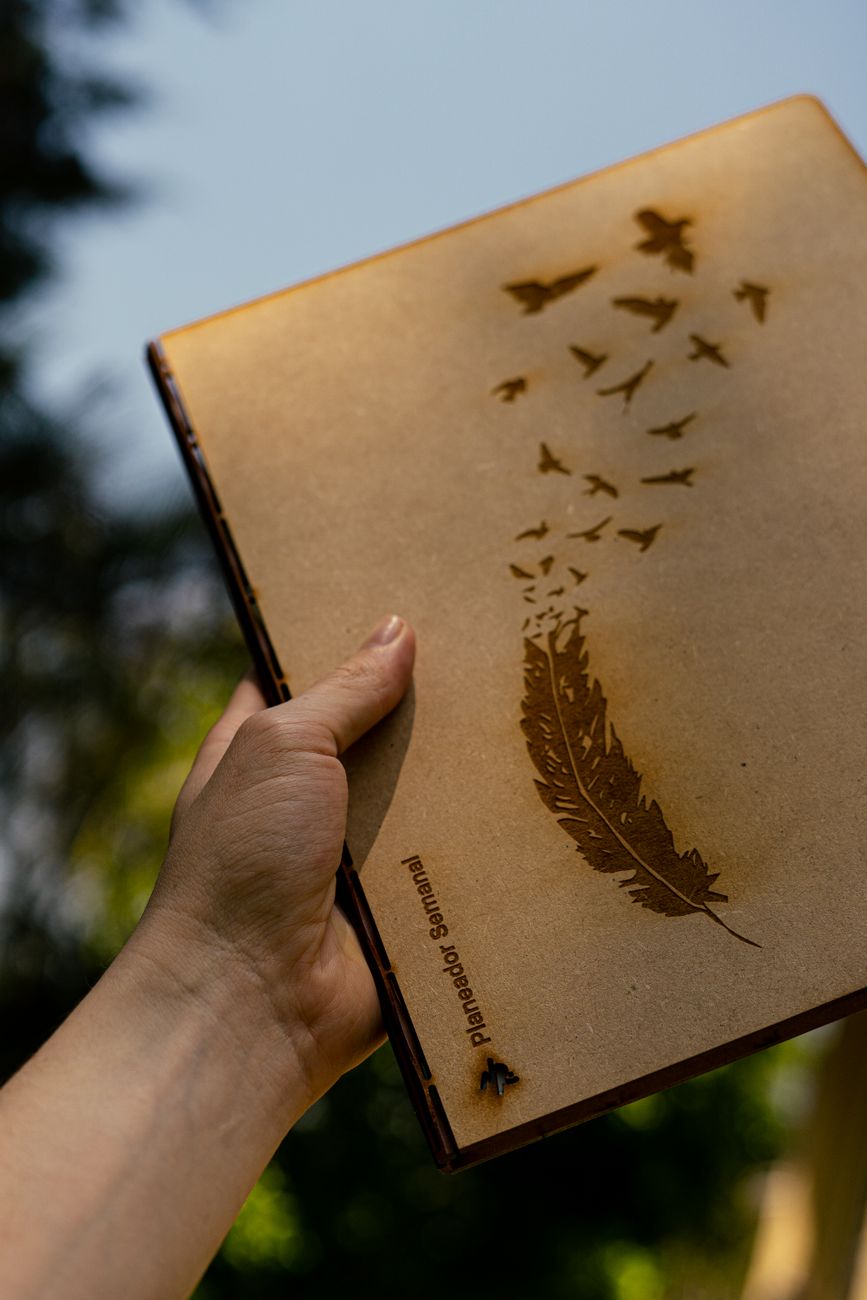 Close-up of a hand holding a notebook featuring an elegant feather and bird design in natural outdoor setting.
